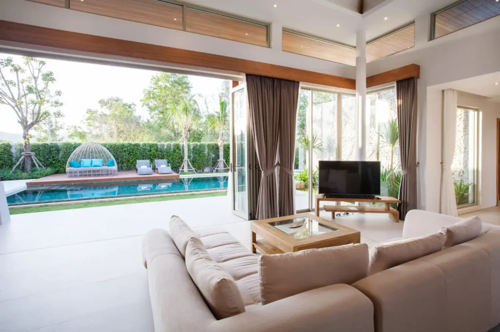Premier villa interior designing services in Dubai by RCi Red Chillies Interiors LLC, featuring a modern living room with large windows overlooking a beautiful pool and garden area, stylish beige sofa, and wooden furniture
