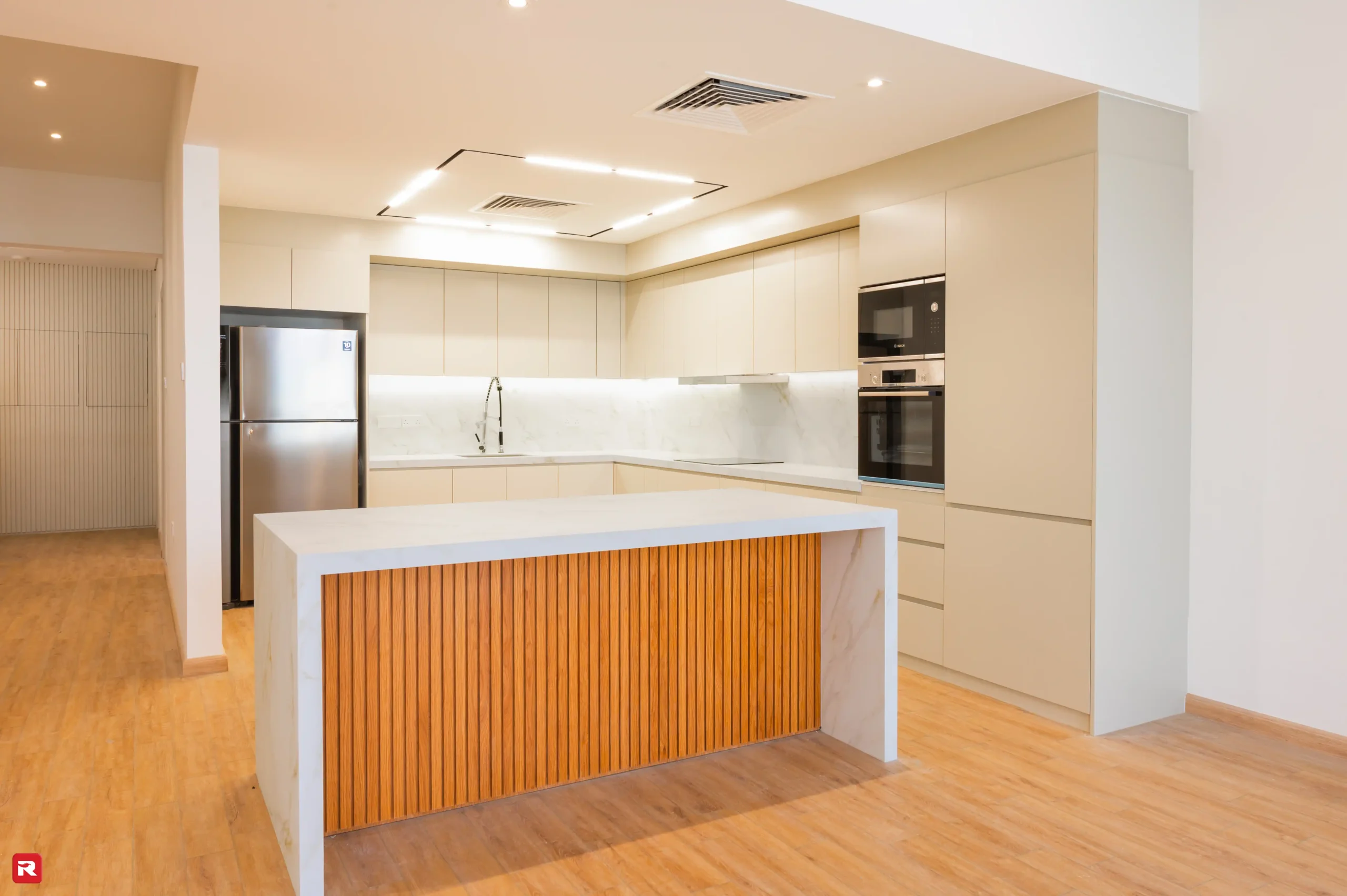 Modern kitchen with beige cabinets, stainless steel appliances, white marble countertop island with wooden slat accents in a villa at Al Furjan by RCi-Red Chillies Interiors